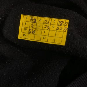 K&ouml;nnte beinhalten: Nahaufnahme eines schwarzen Stoffes mit einem gelben Etikett. Das Etikett enth&auml;lt handschriftliche Zahlen und Buchstaben in schwarzer Tinte. Der Stoff scheint ein dunkles Material zu sein, m&ouml;glicherweise Wolle oder Baumwolle.