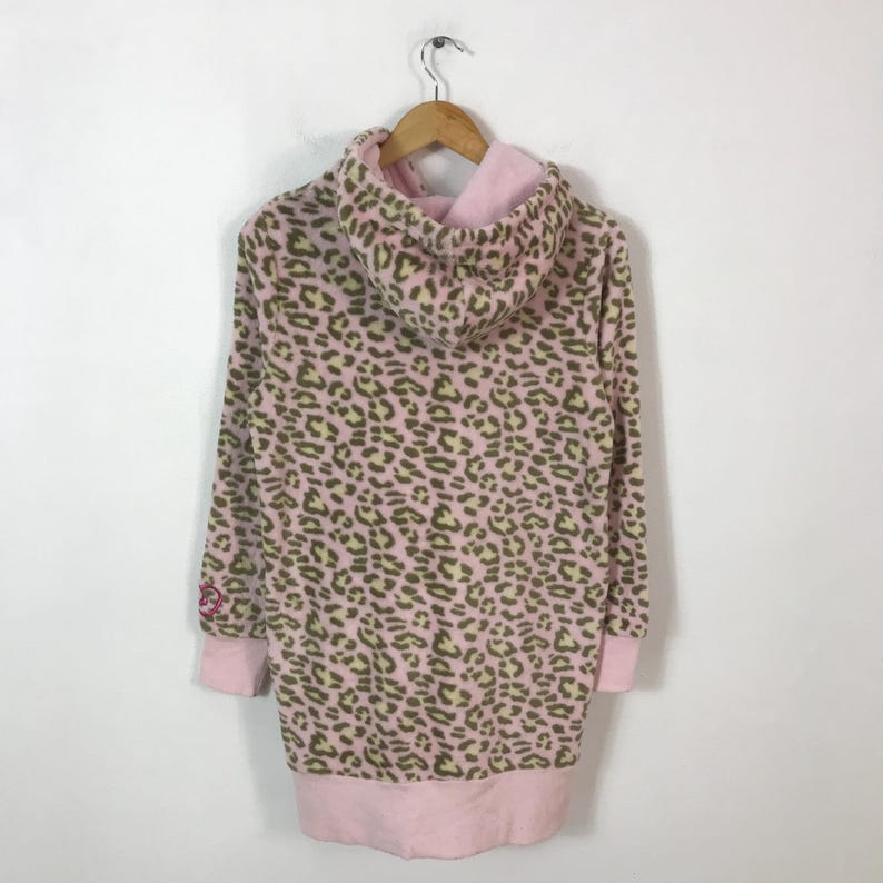 Puede incluir: Una sudadera con capucha rosa con un estampado de leopardo en marr&oacute;n y verde claro. La sudadera tiene mangas largas y un forro de capucha y pu&ntilde;os rosas. La prenda est&aacute; colgada de una percha de madera.