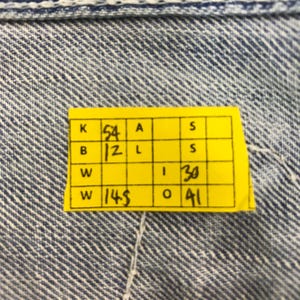 May include: Close-up of a yellow tag with black text and numbers, attached to blue denim fabric. The tag has several boxes with numbers including 54, 12, 30, 41, and 145. The denim has a textured, woven pattern.