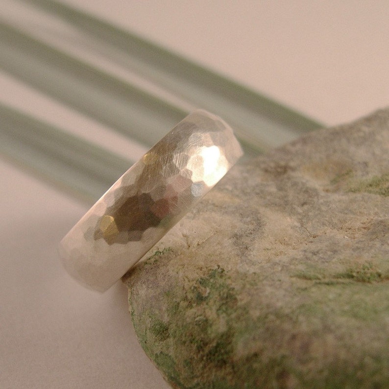 May include: A silver hammered wedding band ring, with a textured surface, resting on a gray and green rock.