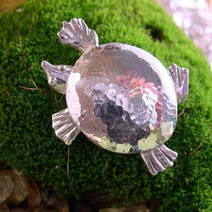 Puede incluir: Un broche de plata en forma de tortuga con un acabado martillado. La tortuga está mirando hacia la derecha y tiene un caparazón detallado.