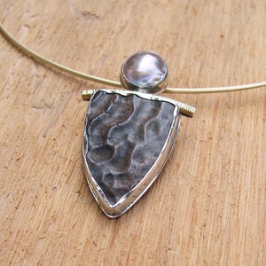 May include: A silver pendant necklace with a textured, dark brown stone set in a silver frame. The stone is surrounded by a gold trim. The pendant is attached to a thin silver chain.