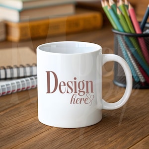 Puede incluir: Una taza de cerámica blanca con un asa curvada. La taza presenta las palabras "Design here" en una fuente marrón. La taza está sobre una superficie de madera, con lápices y cuadernos en el fondo.
