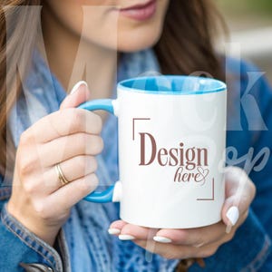 May include: A white ceramic mug with a blue rim, featuring the text "Design here" in brown lettering. The mug is being held by a person.