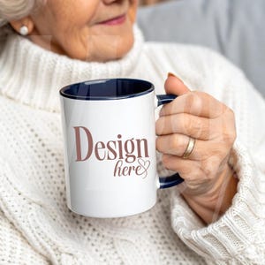 May include: White ceramic mug with a navy blue interior and handle. The mug features the text "Design here" in a brown script font. The mug is held by a person wearing a white sweater.