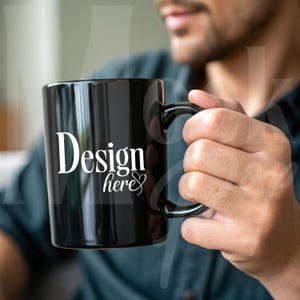 May include: A black ceramic mug with a white design that says "Design here" with a heart symbol. The mug is being held by a person.