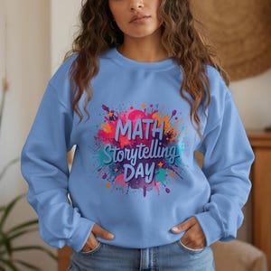 May include: A light blue sweatshirt with the words "MATH Storytelling DAY" in a colorful, splash-like design. The sweatshirt is a crew neck style and has long sleeves. The overall theme is education and learning.