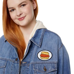 Pode incluir: Casaco de ganga azul claro com um patch com um sanduíche de frango de desenho animado. O patch é oval, com fundo branco e borda preta. O casaco tem um design clássico com fecho de botão e bolsos.