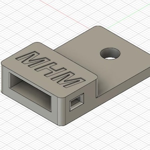 Puede incluir: Objeto gris claro impreso en 3D con las letras "MHM" grabadas en la superficie superior. Tiene una base rectangular con un recorte, un lado rectangular más pequeño y una plataforma elevada con un agujero circular.