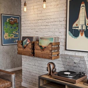 May include: Two dark brown wooden wall-mounted crates holding vinyl records, positioned above a wooden media console with a turntable and headphones. The wall is white brick, flanked by framed posters of a pixelated map and a space shuttle launch.