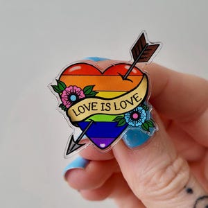 Puede incluir: Un pin en forma de corazón con un diseño de gradiente de arcoíris, atravesado por una flecha. El pin presenta una pancarta con el texto "LOVE IS LOVE" y está adornado con detalles florales. El pin es sostenido por una persona con esmalte de uñas azul.
