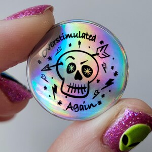 May include: A holographic button with a black and white skull graphic and the text "Overstimulated Again". The skull has an arrow through it and is surrounded by stars and lightning bolts.