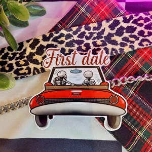 May include: A red car sticker with two skeletons inside, driving on a first date. The sticker says "First date" in a white font.