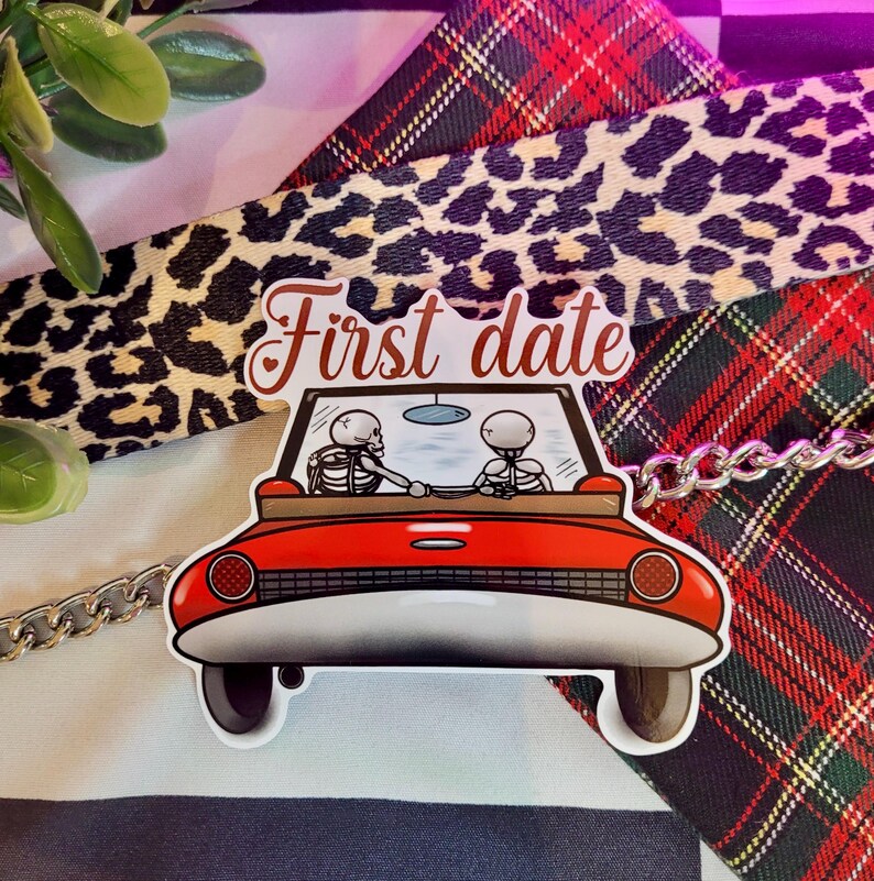 May include: A red car sticker with two skeletons inside, driving on a first date. The sticker says "First date" in a white font.