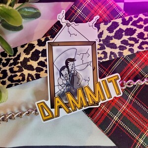 May include: A yellow sticker with the word "DAMMIT" in bold letters. The sticker features a cartoon image of a broken picture frame with two people inside. The sticker is on a red and black plaid fabric.