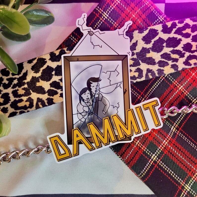 May include: A yellow sticker with the word "DAMMIT" in bold letters. The sticker features a cartoon image of a broken picture frame with two people inside. The sticker is on a red and black plaid fabric.