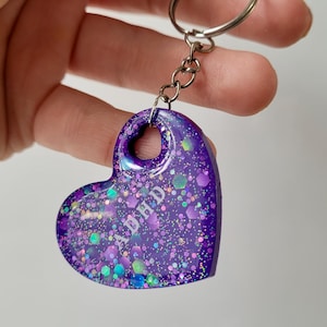 Può includere: Un portachiavi a forma di cuore viola con glitter e le lettere "ADHD". Il portachiavi è attaccato a un anello e una catena di metallo argentato. Il cuore ha un foro nella parte superiore ed è tenuto da una mano su sfondo bianco.