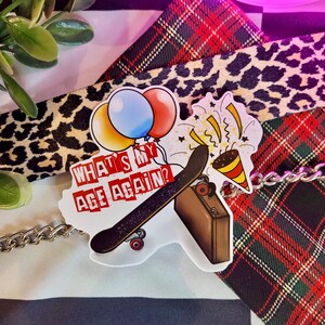 May include: A white sticker with a skateboard, a suitcase, party hats, and balloons. The text on the sticker reads "What's my age again?"