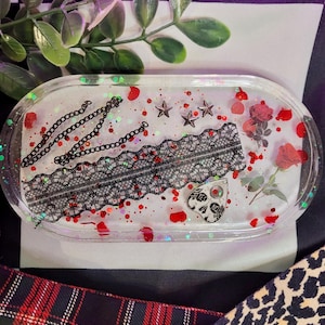 Op de afbeelding: Een transparante harsbak met een zwart kantontwerp, rode glitter en zwarte kettingen. De bak heeft ook een klein zwart-wit Ouija-bordbedeltje, rode en witte bloemen en zilveren sterren.