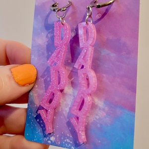 May include: Two pink glitter earrings spelling "DADDY" vertically. The earrings hang from silver-coloured ear wires, presented against a purple and blue backdrop. The earrings are a fun, playful accessory.