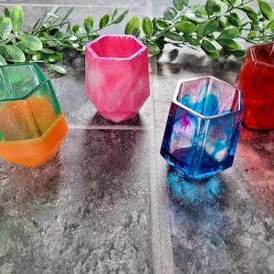 Puede incluir: Cuatro vasos hexagonales de resina en diferentes colores: verde y naranja, rosa, azul con remolinos blancos y rojo. Los vasos están sobre una superficie de baldosas grises.