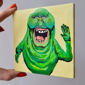 Puede incluir: Una pequeña pintura sobre lienzo cuadrado que representa un personaje de dibujos animados verde con la boca abierta y una mano saludando. El personaje tiene ojos y dientes rosados. El fondo es de color amarillo claro.