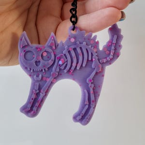 May include: A purple cat skeleton pendant with pink glitter accents. The cat-shaped charm has a detailed skeletal design and a black chain. The pendant is a Halloween-themed accessory.