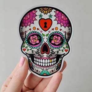Può includere: Un teschio di zucchero colorato con fiori rosa e bianchi, un cuore con un buco della serratura e un dente d'oro. Il teschio è decorato con foglie verdi e vortici blu e gialli.