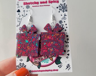 May include: Red glitter gift box earrings with silver hooks. The earrings are handmade and feature a square shape with a bow on top. The earrings are displayed on a white card with the text "Handmade Earrings by Sketchy and Spicy".