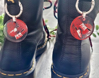Könnte beinhalten: Schwarze Lederstiefel mit gelben Nähten und rot-schwarzen Schnürsenkeln. Jeder Stiefel hat einen silbernen Schlüsselanhänger mit einem runden, roten Anhänger mit weißen Messern.