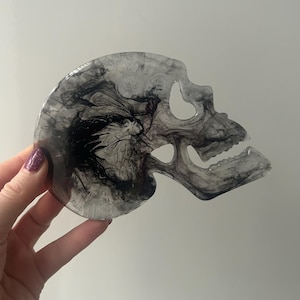 Puede incluir: Un posavasos transparente en forma de calavera con un diseño de tinta negra en espiral. La calavera tiene cuencas de ojos abiertas y una mandíbula detallada. El posavasos está hecho de resina.