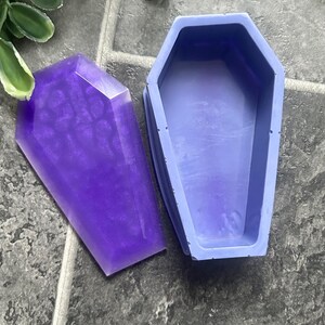 May include: A purple coffin-shaped container with a matching lid. The lid has a shiny, marbled appearance. The container is open, revealing a hollow interior. The items are on a gray tiled surface. The theme is gothic or Halloween.