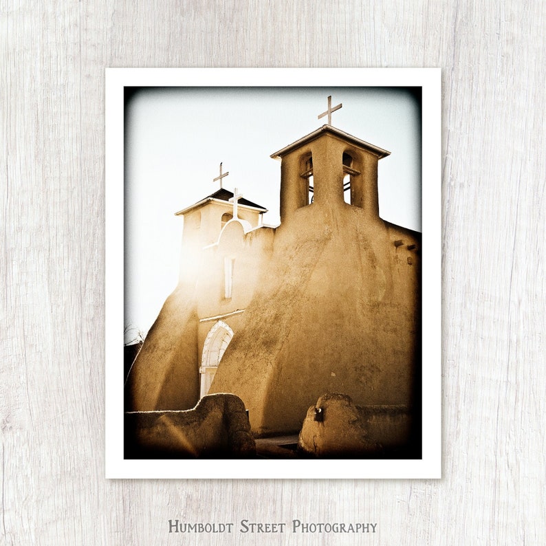 May include: A sepia-toned photograph of a historic adobe church with two bell towers and a cross on top of each tower. The church is surrounded by a white frame.