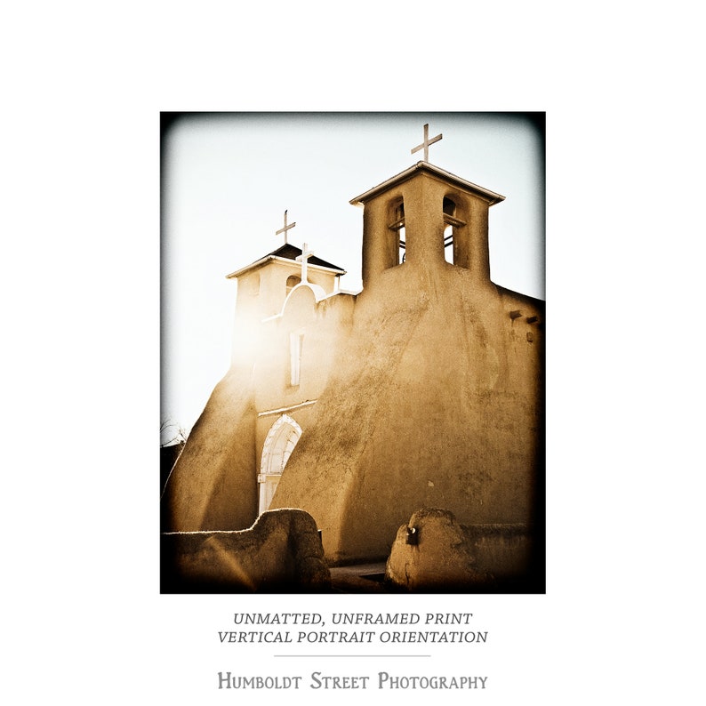 May include: A vintage-style photograph of a tan adobe church with two bell towers and a white cross on top of each tower. The church is lit by the sun, casting a warm glow on the building.