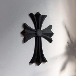 Könnte beinhalten: Eine schwarze, stilisierte Kreuz-Wanddekoration mit einem erhabenen, runden Zentrum. Das Kreuz besteht aus einem glatten, harten Material und hat ein modernes, minimalistisches Design.