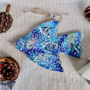 Peut inclure: Ornement en forme d'oiseau bleu peint à la main, avec des motifs floraux blancs, verts et violets. Il est suspendu à une ficelle. Des pommes de pin et un sapin de Noël décoratif suggèrent un thème de vacances.