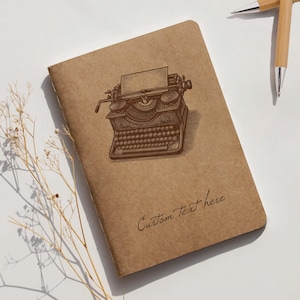 以下が含まれることがあります： ヴィンテージのタイプライターのイラストと「Custom Text here」の文字が入った茶色のノート。背景には2本のペンが見えます。ノートは長方形で、柔らかく、質感のある表紙が付いています。