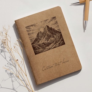 Op de afbeelding: Een bruin notitieboek met een berglandschap illustratie en de tekst "Custom Text here". Twee bamboe pennen liggen rechts. Het notitieboek ligt op een wit oppervlak.