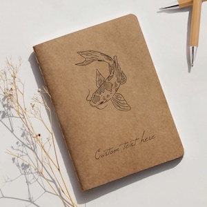 Könnte beinhalten: Ein braunes Notizbuch mit einer Koi-Fisch-Illustration und dem Text "Custom Text here". Zwei Stifte aus Bambus liegen auf der weißen Oberfläche. Ein Zweig getrockneter Blumen befindet sich in der Ecke.