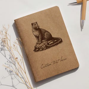 May include: A brown notebook featuring a detailed illustration of a leopard on a rock. The text "Custom Text here" is written in cursive. Two bamboo pens are visible in the background.