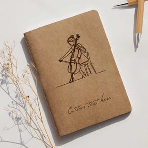 May include: A brown notebook featuring a line drawing of a cellist. The words "Custom Text here" are written in cursive. Two bamboo pens are visible in the background. The notebook is on a white surface.