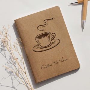 May include: A brown notebook with a coffee cup illustration and the text "Custom Text here". The notebook is lying on a white surface with two pens and a sprig of dried flowers.