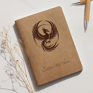 May include: A brown notebook with a phoenix design in the center and the words "Custom Text here" at the bottom. Two bamboo pens are in the upper right corner. A sprig of dried flowers is in the lower left corner.