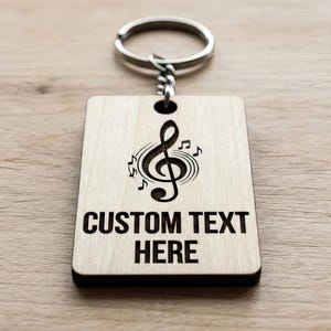 Op de afbeelding: Een houten sleutelhanger met een zilveren ring. De rechthoekige tag heeft een ontwerp van een G-sleutel en muzieknoten, met de woorden "CUSTOM TEXT HERE" in vette, zwarte letters. De sleutelhanger ligt op een licht houten oppervlak.