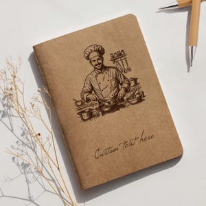 Op de afbeelding: Een bruin notitieboek met een chef-illustratie en de tekst "Custom Text here". De illustratie toont een kok die aan het koken is. Twee bamboe pennen zijn ook zichtbaar.