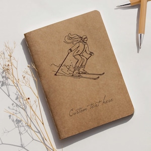 Könnte beinhalten: Ein braunes Notizbuch mit einer Skifahrer-Illustration und den Worten "Custom Text here". Zwei Stifte aus Bambus befinden sich im Hintergrund. Das Notizbuch ist etwa 14 cm hoch und 10 cm breit.