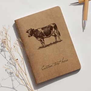 Peut inclure: Un carnet marron avec une illustration de vache et le texte "Custom Text here". La vache est marron et blanche, dessinée dans un style esquisse détaillé. Deux stylos en bambou sont en arrière-plan. Le carnet mesure environ 21 cm de long.