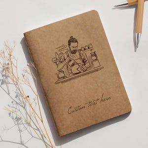 Peut inclure: Un carnet marron avec une illustration détaillée d'un barista versant du café. Le carnet porte l'inscription "Custom Text here" en écriture cursive. Deux stylos en bambou sont visibles.
