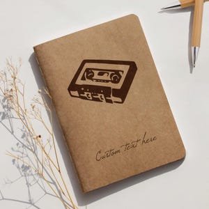 May include: A brown notebook with a retro cassette tape design. The notebook has the words "Custom Text here" printed on the cover. Two bamboo pens are in the background.