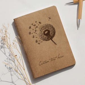 May include: A brown notebook with a dandelion design and the text "Custom Text here." The notebook is rectangular and has a soft cover. Two pens are visible in the background. The notebook is a stationery item.